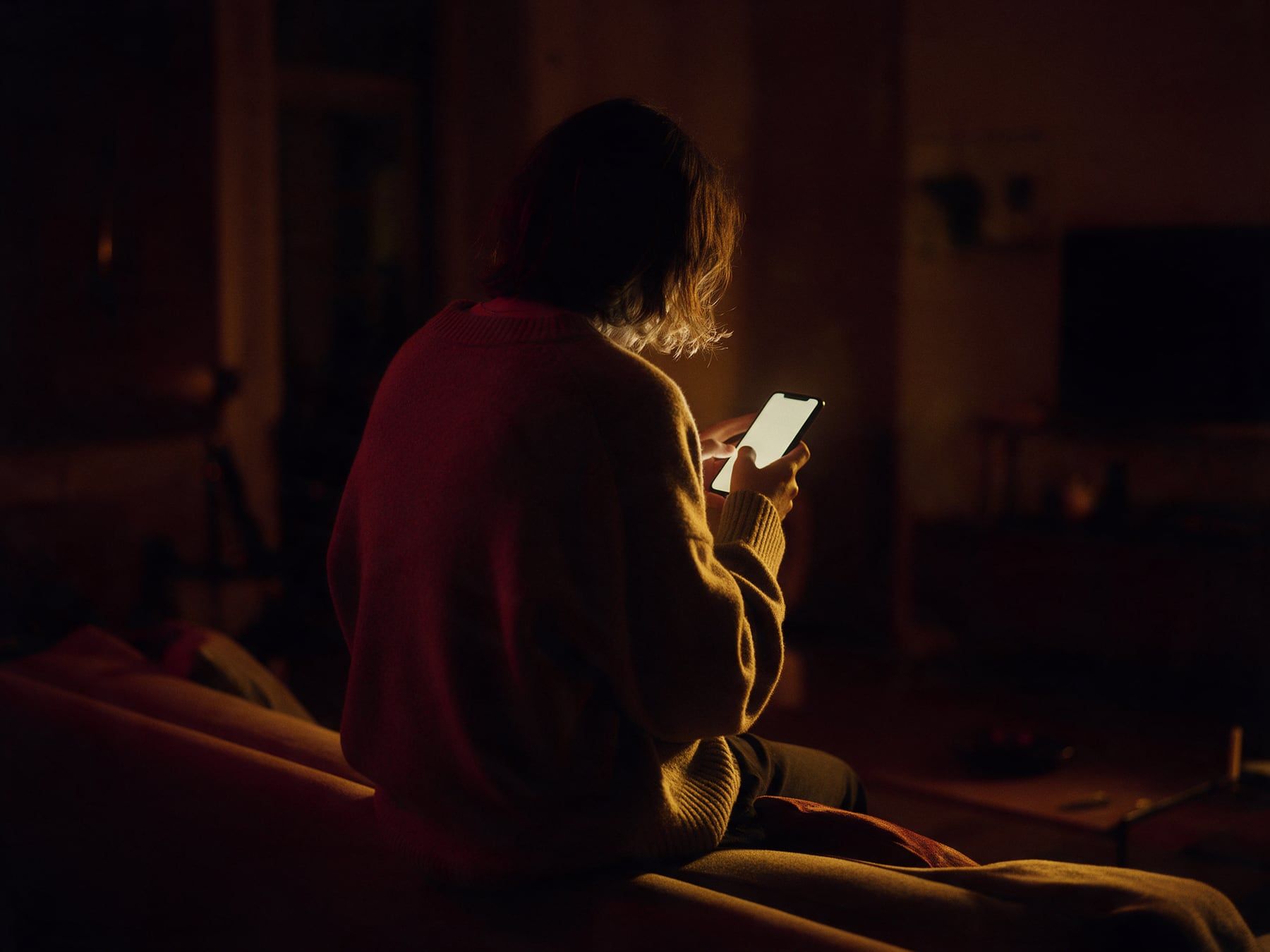 Une personne de dos, assise sur un canapé, lit son téléphone le soir