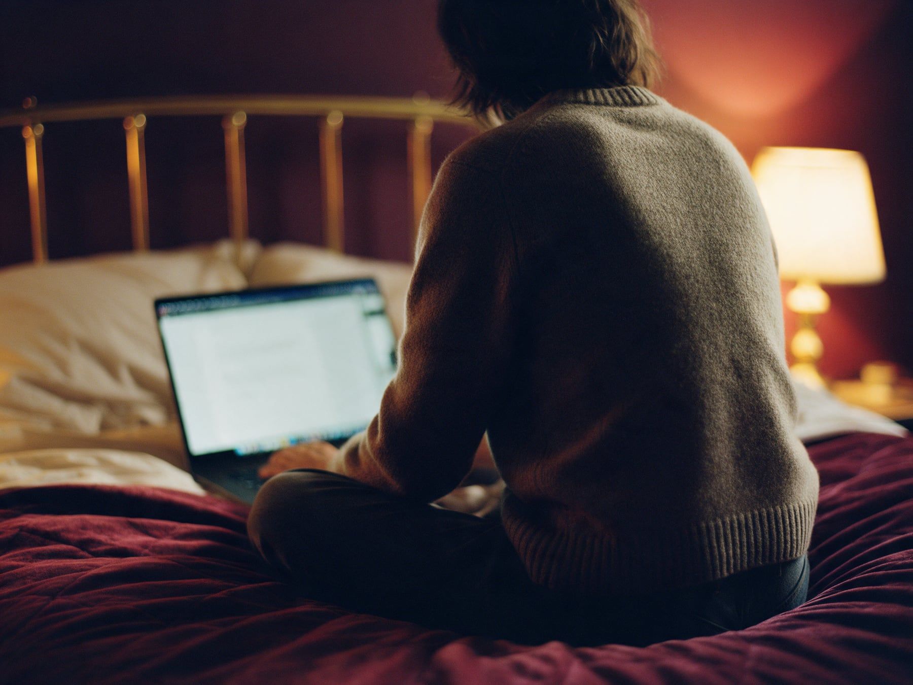 Une personne assise sur un lit consulte un ordinateur portable le soir